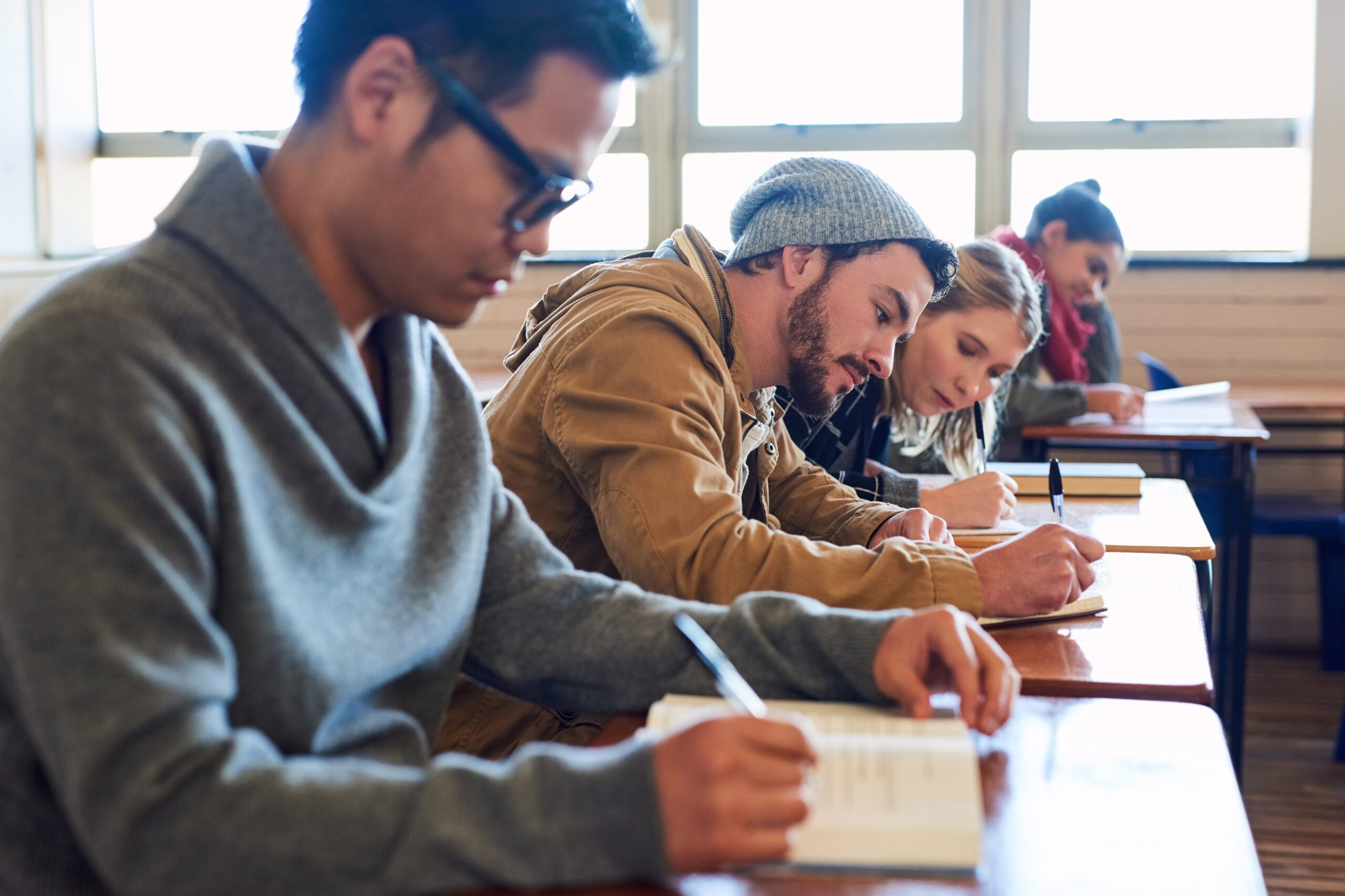 College students, writing and desk in classroom with books for english literature or language for exam. People, university and scholarship for learning or education, knowledge and academic work