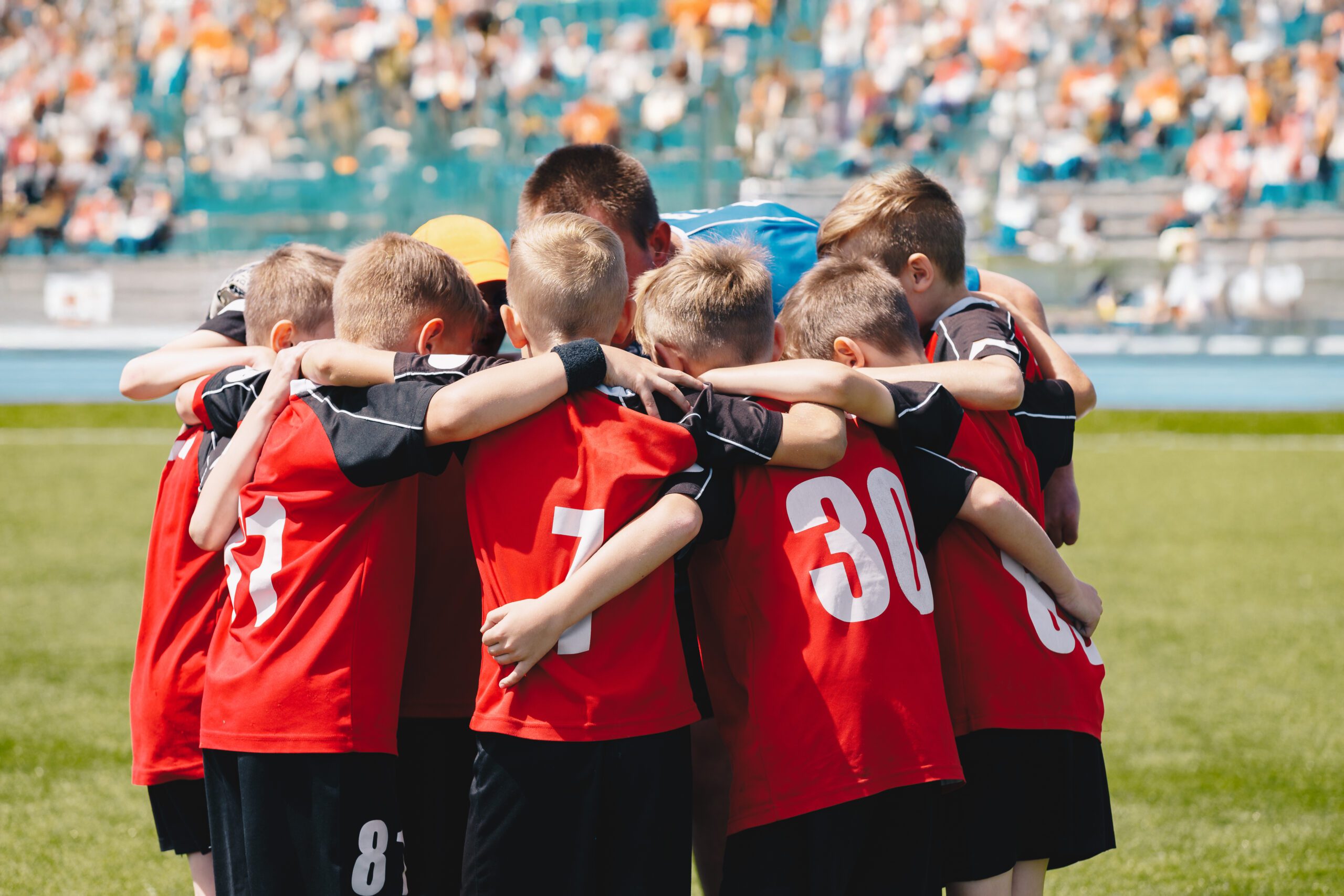 Teenage boys in a sports team. Sports team friendship. Group of sports players with coach huddling in a circle and motivating each other. Boys in red jersey shirts with numbers